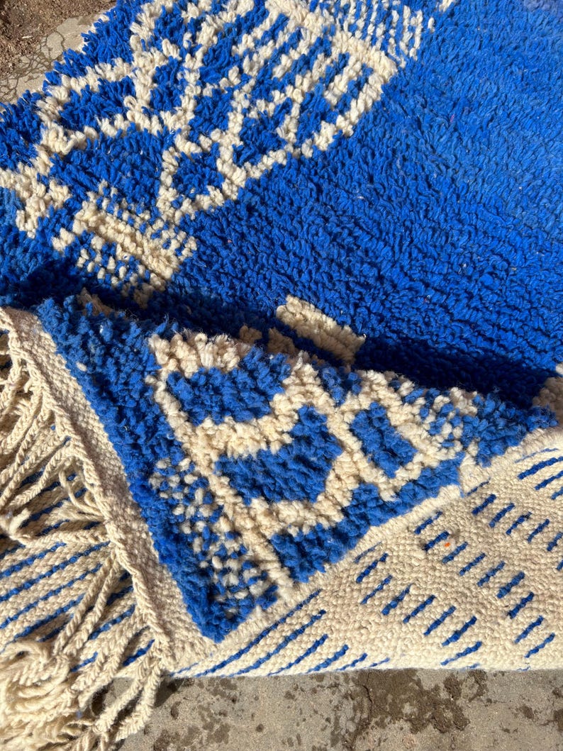 Op de afbeelding: Een blauw en wit Marokkaans tapijt met een geometrisch patroon en franjes. Het tapijt is gemaakt van wol en heeft een zachte, pluche textuur.