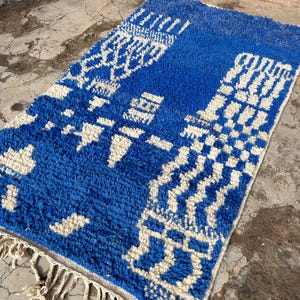 Op de afbeelding: Een blauw en wit Marokkaans tapijt met een geometrisch patroon. Het tapijt heeft een franje rand en is gemaakt van wol.