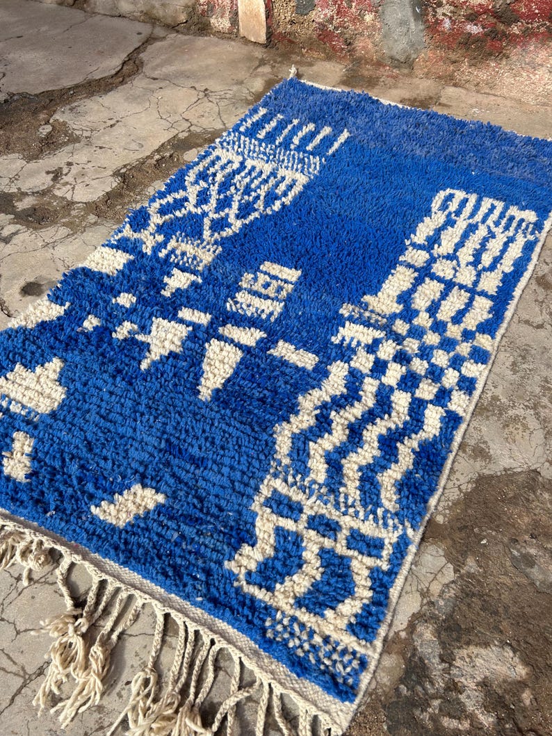 Op de afbeelding: Een blauw en wit Marokkaans tapijt met een geometrisch patroon. Het tapijt heeft een franje rand en is gemaakt van wol.