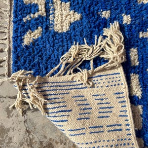 Op de afbeelding: Een blauw en wit geweven tapijt met franjes aan alle kanten. Het tapijt heeft een geometrisch patroon en is doormidden gevouwen.
