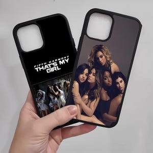 May include: Two black phone cases. One features the text "FIFTH HARMONY THAT'S MY GIRL" in white, with a photo of the group. The other case displays a photo of the Fifth Harmony group. The cases are held in a hand.