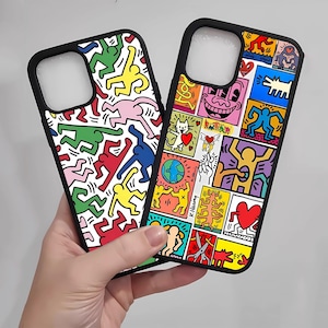 May include: Two black phone cases with colorful pop art designs. One case features repeating figures in red, green, yellow, pink, and blue on a white background. The other case has a collage of images, including a smiling face, a dog, and a heart.