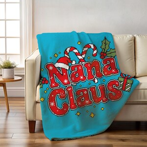 May include: A turquoise knitted throw blanket with the words "Nana Claus" in red pixelated letters. The blanket features Christmas-themed graphics like candy canes, a Santa hat, holly, and a wrapped gift. The blanket is draped over a beige sofa.