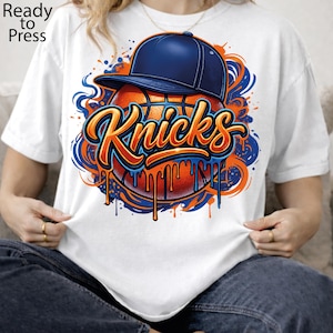 May include: A white t-shirt featuring a graphic of a basketball wearing a dark blue baseball cap. The word "Knicks" is written in orange script across the center, with blue and orange paint splatter effects dripping down the basketball design.