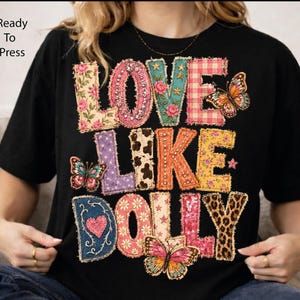 May include: A black t-shirt featuring the text LOVE LIKE DOLLY in large, multi-patterned letters. The design includes floral, gingham, leopard, and cow prints, along with butterfly graphics, sequins, and rhinestones. The text Ready To Press is visible.