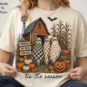 May include: A cream-colored t-shirt featuring a graphic of a red barn, a windmill, corn stalks, and three ghosts with checkered, floral, and polka dot patterns. A sign reads PUMPKINS, HAY-RIDES, CORN MAZE. Text at the bottom reads tis the season.