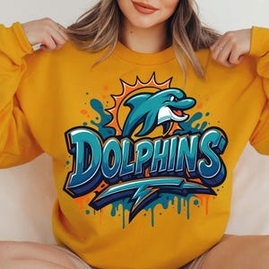 Może przedstawiać: Musztardowa ż&oacute;łta bluza z turkusowym delfinem wyskakującym z wody i napisem "Dolphins" w kolorze turkusowym i białym. Projekt zawiera pomarańczowe akcenty i błyskawicę. Swobodny, sportowy styl.