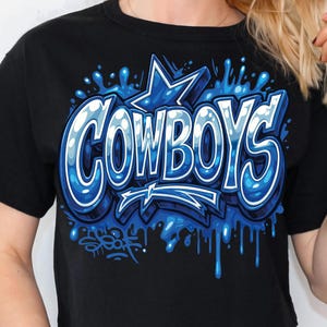 Może przedstawiać: Czarny t-shirt z napisem "COWBOYS" w niebieskim stylu graffiti. Słowo jest obrysowane na biało z niebieską gwiazdą i kroplami niebieskiej farby. Koszulka ma kr&oacute;tkie rękawy.