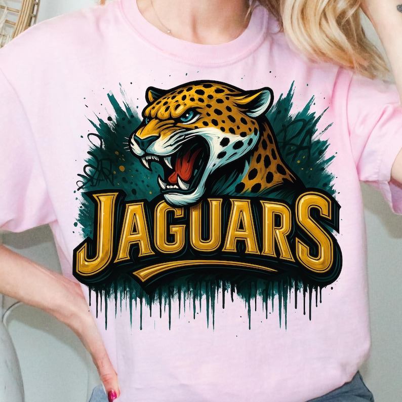 Może przedstawiać: R&oacute;żowa koszulka z grafiką jaguara z otwartą paszczą, ż&oacute;łtymi oczami i czarnymi plamami. Napis "JAGUARS" jest w złotych literach na ciemnozielonym tle.