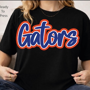 May include: A black short-sleeved t-shirt featuring the word Gators in a blue script font with a thick orange outline. The text Ready To Press is printed in the upper left corner. The shirt has a crew neckline and a standard fit design.