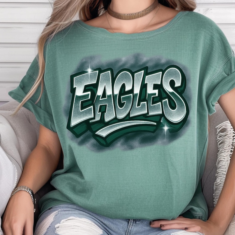 K&ouml;nnte beinhalten: Ein gr&uuml;nes T-Shirt mit dem Wort "EAGLES" in einem wei&szlig;en und gr&uuml;nen 3D-Design. Das Shirt hat hochgekrempelte &Auml;rmel und einen Rundhalsausschnitt. Der Text ist wei&szlig; umrandet mit einem gr&uuml;nen Schatten und einem dunkelgr&uuml;nen Hintergrund.