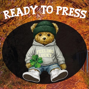 May include: A teddy bear illustration wearing a green beanie, a gray hoodie with "FEELING LUCKY" text, blue jeans, and white sneakers, holding a four-leaf clover. The image has the text "READY TO PRESS" at the top.