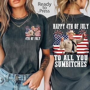 May include: A charcoal grey t-shirt featuring a graphic of a man in a tan uniform and cowboy hat standing before an American flag with fireworks. The front has a small chest print, while the back displays the text Happy 4th of July to all you sumbitches.
