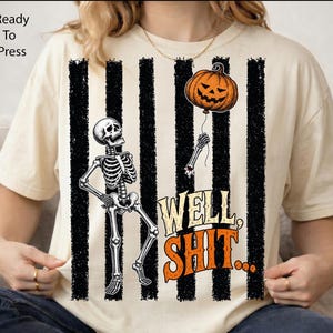 May include: A cream-colored t-shirt featuring a graphic of a skeleton holding a string attached to an orange jack-o'-lantern balloon. The background has vertical black stripes, and the text reads, "WELL, SHIT..." in orange and cream letters.