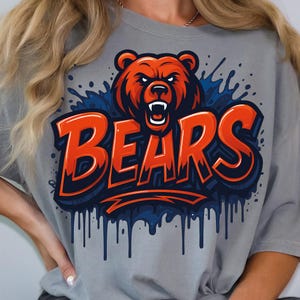 Może przedstawiać: Szara koszulka z efektowną grafiką niedźwiedzia w odcieniach pomarańczu i czerwieni, z napisem "BEARS" pogrubioną pomarańczową czcionką. Projekt zawiera niebieskie akcenty i efekty kapania, tworząc dynamiczny wygląd. Casualowy i sportowy element odzieży.