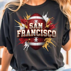 May include: Black t-shirt featuring a football design with the words "SAN FRANCISCO" in a 3D effect. The football is red and white, with gold, red, and white paint splatter accents. A sporty graphic tee.