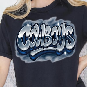 K&ouml;nnte beinhalten: Marineblaues T-Shirt mit dem Wort "COWBOYS" in einem silbernen, blauen und wei&szlig;en 3D-Design. Der Text ist vor einem rauchigen, wolkenartigen Hintergrund platziert. Das Shirt hat kurze &Auml;rmel.