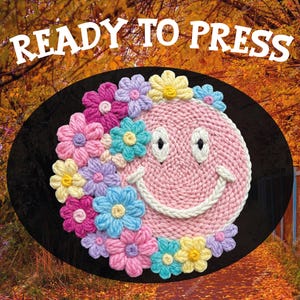 May include: A pink, knitted smiley face with a white stitched smile and eyes, surrounded by colorful crocheted flowers. The text "READY TO PRESS" is displayed above the smiley face on a black oval.
