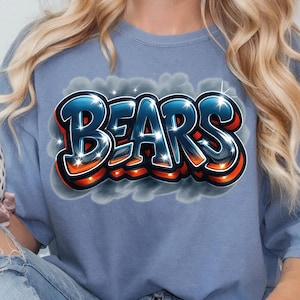 K&ouml;nnte beinhalten: Ein blaues T-Shirt mit dem Wort "BEARS" im Graffiti-Stil. Der Text hat eine schwarze Umrandung, orangefarbene und wei&szlig;e Akzente und einen blauen Farbverlauf. Das Design befindet sich vor einem Hintergrund aus grauen Wolken.