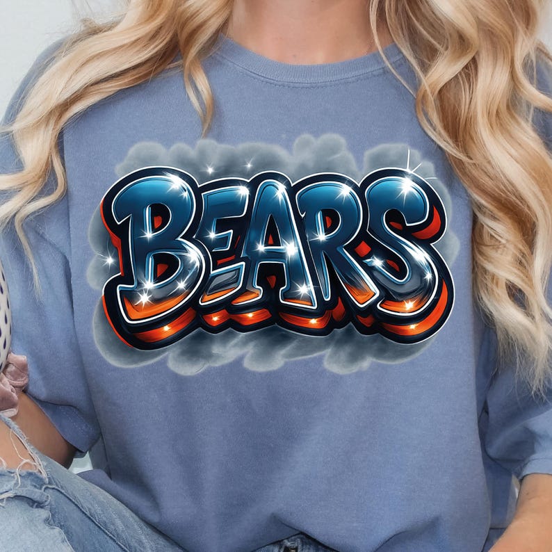 K&ouml;nnte beinhalten: Ein blaues T-Shirt mit dem Wort "BEARS" im Graffiti-Stil. Der Text hat eine schwarze Umrandung, orangefarbene und wei&szlig;e Akzente und einen blauen Farbverlauf. Das Design befindet sich vor einem Hintergrund aus grauen Wolken.