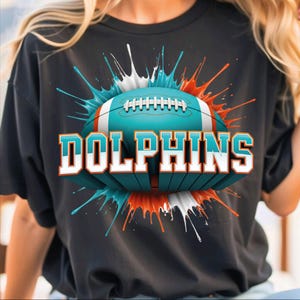 May include: Black t-shirt featuring a teal and white football with the word "DOLPHINS" in teal and white block letters. The football is surrounded by teal, white, and orange paint splatters.