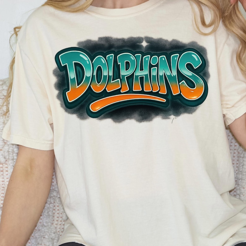 K&ouml;nnte beinhalten: Cremefarbenes T-Shirt mit dem Wort "Dolphins" in einem Graffiti-Design. Der Text ist in Teal, Orange und Wei&szlig; gehalten, schwarz umrandet, vor einem dunklen, grauen, wolkenartigen Hintergrund.