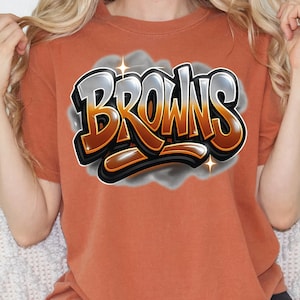 K&ouml;nnte beinhalten: Ein orangefarbenes T-Shirt mit dem Wort "BROWNS" in einem Graffiti-Stil. Der Text ist braun mit silbernen und schwarzen Akzenten, vor einem grauen Wolkenhintergrund mit Sternen.
