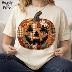 May include: A cream-coloured t-shirt featuring a large, central pumpkin graphic composed of various fabric-style patches in shades of orange, brown, and green with leopard and plaid patterns. The pumpkin has black stitched eyes and a mouth.