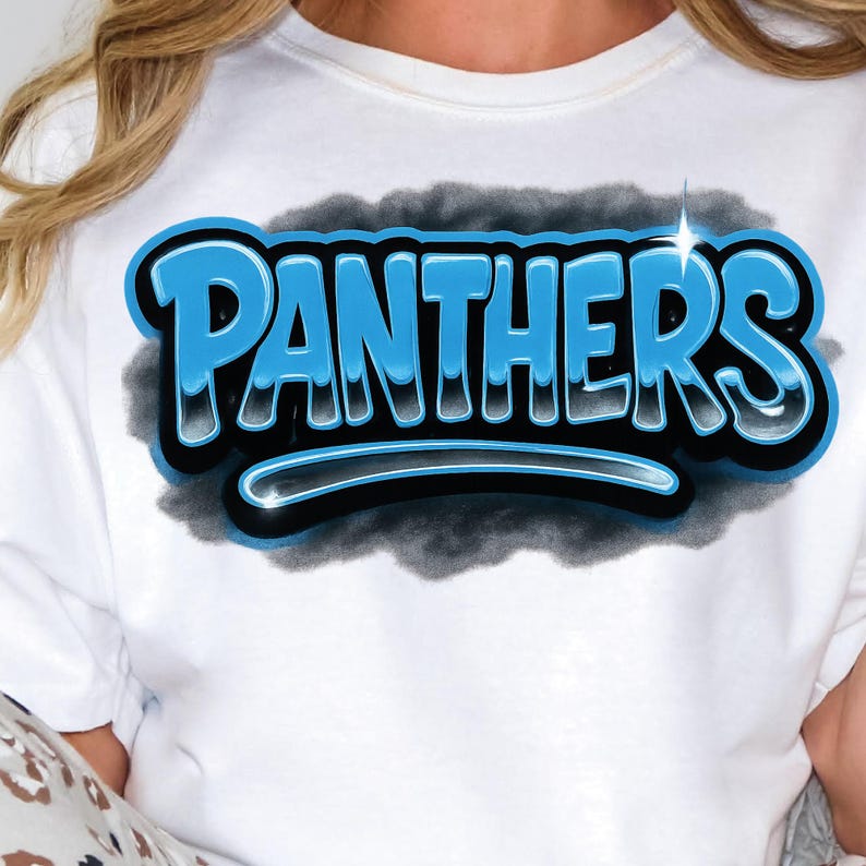 K&ouml;nnte beinhalten: Wei&szlig;es T-Shirt mit dem Wort "PANTHERS" in einem blauen, schwarzen und wei&szlig;en 3D-Design. Das Wort ist vor einem dunkelgrauen, wolkenartigen Hintergrund platziert. Das Shirt hat einen Rundhalsausschnitt.