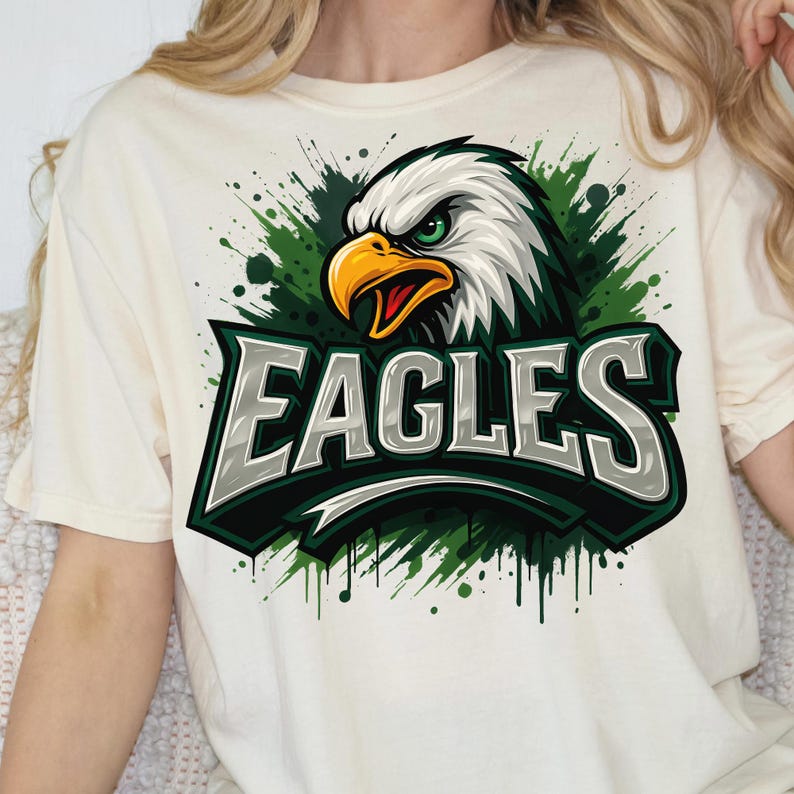 Może przedstawiać: Kremowa koszulka z grafiką orła z zielonymi oczami i ż&oacute;łtym dziobem. Słowo "EAGLES" jest wyświetlane pogrubioną, srebrną czcionką z zielonym obrysem. Projekt zawiera zielone rozpryski.