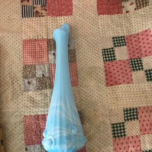 Puede incluir: Un jarr&oacute;n alto de vidrio azul claro con base acampanada y cuerpo esbelto y alargado. El jarr&oacute;n presenta un dise&ntilde;o decorativo, similar a flores, alrededor de la secci&oacute;n media y una parte superior abierta &uacute;nica. El vidrio tiene un sutil patr&oacute;n arremolinado.