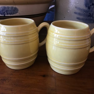 May include: Two yellow ceramic mugs shaped like barrels with white stripes. The mugs have handles and are sitting on a wooden surface.