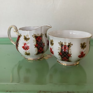 May include: A white porcelain creamer and sugar bowl set with a red and black plaid pattern and gold accents. The design features maple leaves and a gold trim.