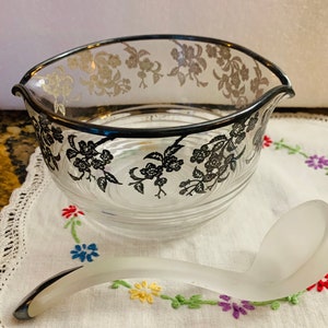 May include: A clear glass serving bowl with a silver rim and floral pattern. The bowl is accompanied by a frosted glass ladle with a silver handle.