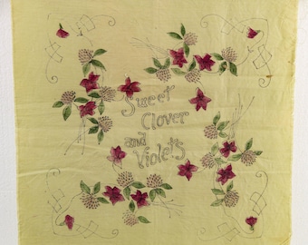 May include: A square, pale yellow fabric with a hand-drawn floral design. The design features red flowers, green leaves, and the words "Sweet Clover and Violets" in cursive script. The fabric is held up by two metal clips.
