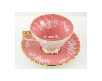 May include: A pink teacup and saucer set with white floral designs and gold trim. The teacup has a scalloped rim and a curved handle. The saucer has a matching design. The set is likely made of porcelain.