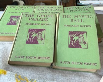 May include: A collection of vintage Judy Bolton mystery books by Margaret Sutton. The books have light green covers with titles like "The Haunted Attic" and "The Mystic Ball." Each book features a purple illustration and text.