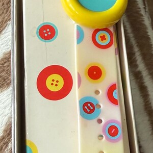 May include: A vintage digital watch with a cream-colored band adorned with colorful circles and button designs. The watch face is yellow with a blue center. The watch is in its original box, which also features button graphics.