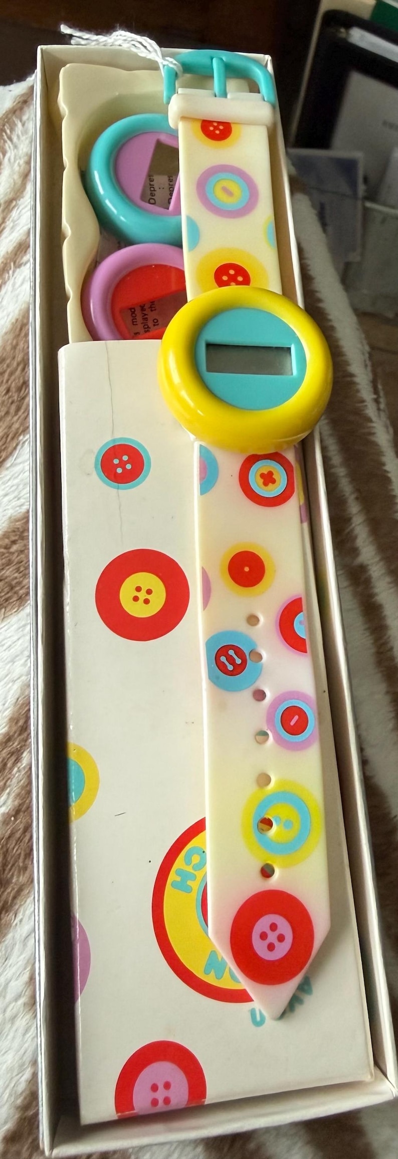 May include: A vintage digital watch with a cream-colored band adorned with colorful circles and button designs. The watch face is yellow with a blue center. The watch is in its original box, which also features button graphics.