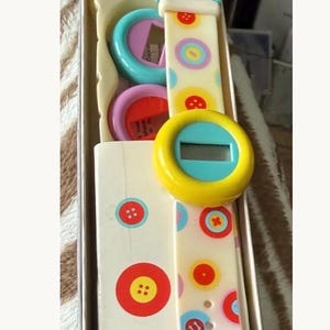 May include: A vintage digital watch with a white strap adorned with colorful button graphics. The watch face is yellow with a blue border and a digital display. The watch is packaged in a box with matching button designs.
