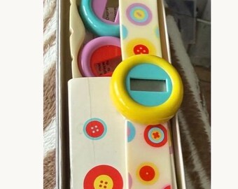 May include: A vintage digital watch with a white strap adorned with colorful button graphics. The watch face is yellow with a blue border and a digital display. The watch is packaged in a box with matching button designs.
