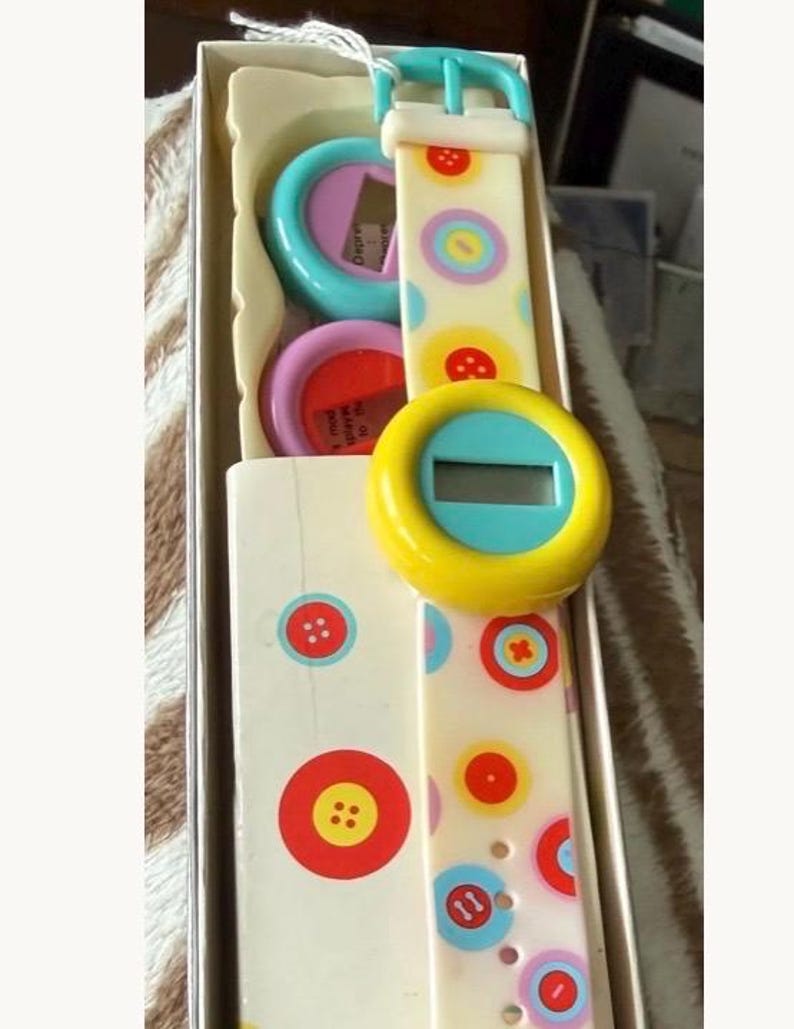May include: A vintage digital watch with a white strap adorned with colorful button graphics. The watch face is yellow with a blue border and a digital display. The watch is packaged in a box with matching button designs.