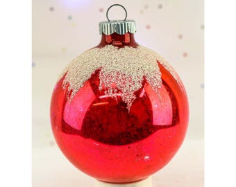 May include: A shiny red Christmas ornament with a silver glitter design on the top. The ornament has a silver metal cap and a small loop for hanging. The background is a soft white with colorful dots.