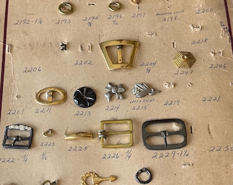 May include: A vintage F. Ronci Co. display board featuring an assortment of metal shoe buckles and ornaments in various shapes, sizes, and finishes. The tan cardboard backing includes printed company information and handwritten reference numbers.