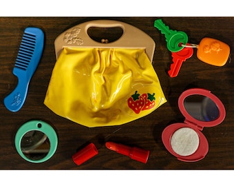 May include: A collection of children's toy accessories, including a yellow purse with a strawberry design, a blue comb, red lipstick, a compact mirror, a small round mirror, and keys.
