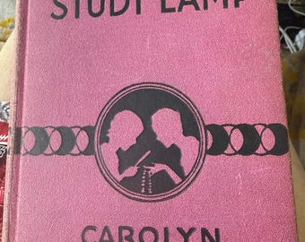 May include: A vintage book titled "By the Light of the Study Lamp" by Carolyn Keene. The cover is pink with black lettering and a circular illustration of two figures. The book's cover is slightly worn.