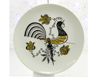 May include: A white ceramic plate with a black and gold rooster design. The rooster is stylized with a large, curved tail and a detailed head. Floral accents and clock hands complete the design. A decorative wall clock.