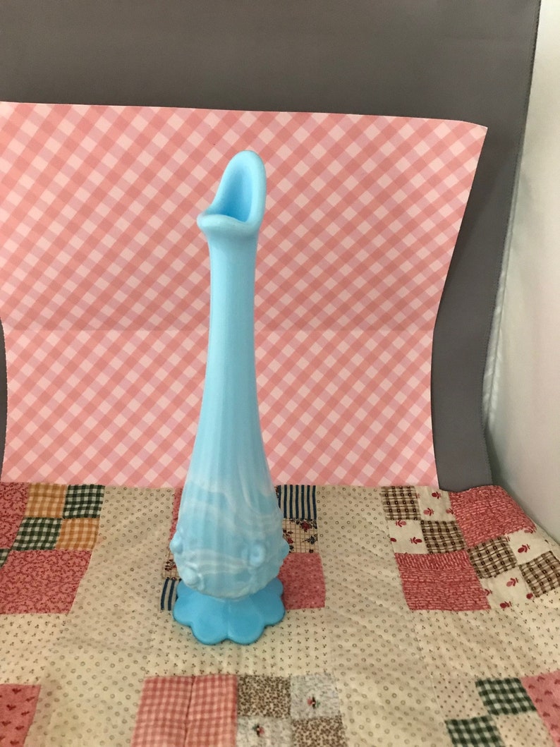 Puede incluir: Un jarr&oacute;n alto de vidrio azul claro con una base acampanada y festoneada y un cuerpo delgado y alargado. El jarr&oacute;n tiene una parte superior &uacute;nica con boca abierta y detalles decorativos en relieve. El vidrio tiene un sutil efecto veteado.