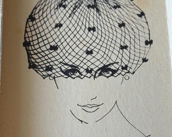 May include: A tan paper card featuring a black ink line drawing of a face wearing a netted veil hat with small accents. The text at the top reads The Famous Glamour Veil Hat, and the bottom right corner includes the printed text NO: followed by a line.