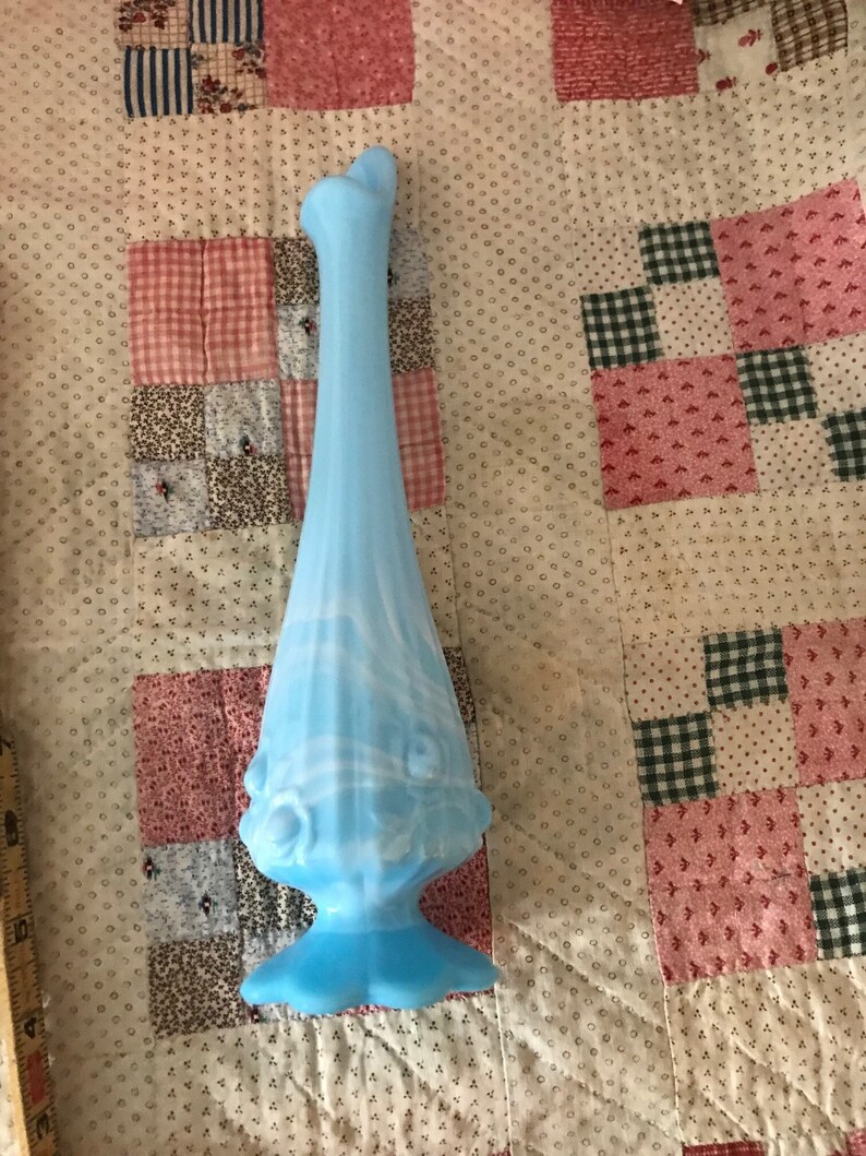 Puede incluir: Un jarr&oacute;n alto de vidrio azul claro con base acampanada y cuerpo esbelto y alargado. El jarr&oacute;n presenta un dise&ntilde;o decorativo, similar a flores, alrededor de la secci&oacute;n media y una parte superior abierta &uacute;nica. El vidrio tiene un sutil patr&oacute;n arremolinado.